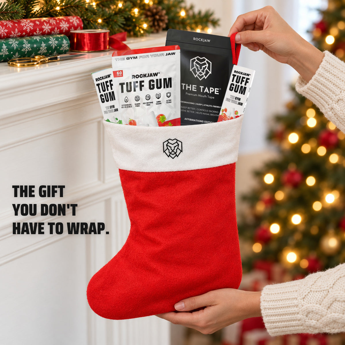 The gift you don't have to wrap. Hands lifting the filled ROCKJAW Stocking by its loop in front of a white mantel on Christmas morning, with rolls of wrapping paper, ribbon and scissors left unused.